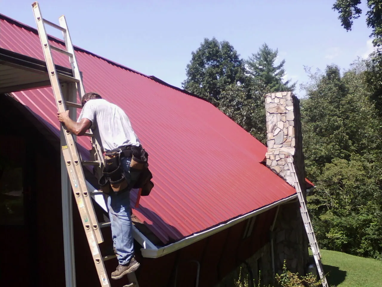 Roofer on a ladder beside a red metal roof and stone chimney — Soffit & Fascia Repair by Roofing Brothers in Speedwell
